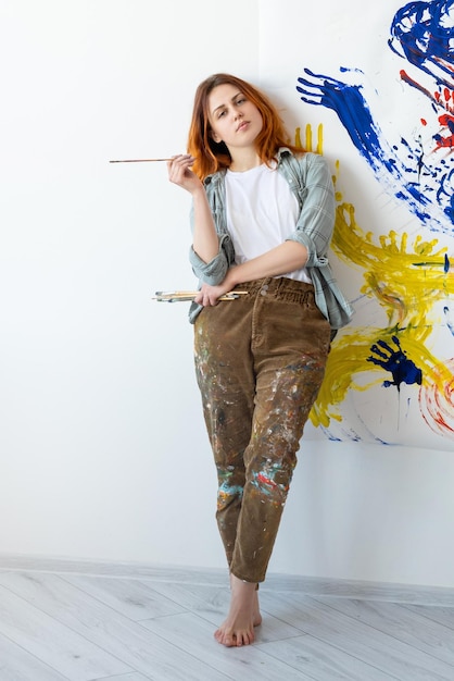 Malowanie ręczne Arteterapia Relaksujący wypoczynek Pewna, utalentowana artystka z pędzlami stojąca przy kolorowej abstrakcyjnej grafice na białej ścianie w galerii światła