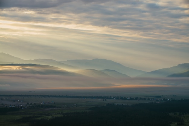 Malowniczy krajobraz górski z pomarańczowym liliowym niskimi chmurami nad wioską wśród sylwetek gór pod zachmurzonym niebem świt. Klimatyczna alpejska sceneria wsi w niskich chmurach w złotym kolorze zachodu słońca.