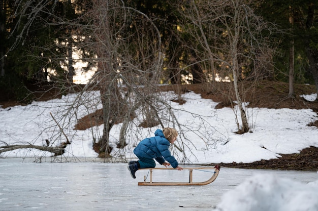 Maluch chłopiec w zimowym garniturze bawi się na zewnątrz w śnieżnej przyrodzie z saniami na naturalnym lodzie