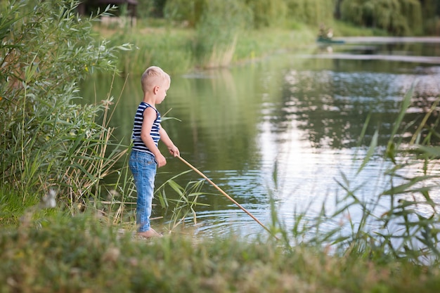 Zdjęcie mały blond chłopiec gra nad rzeką na łodzi. lato zielona trawa i błękitne jezioro.