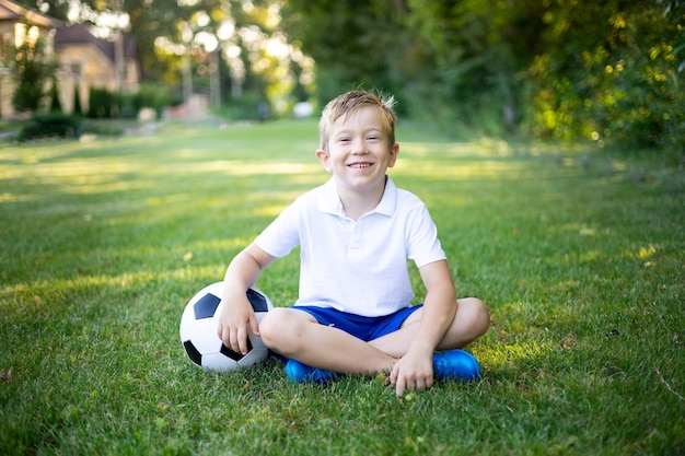 Mały blond chłopiec siedzi latem na trawie z piłką nożną, patrząc w kamerę