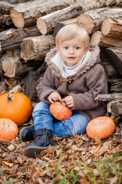 Mały blond chłopiec w jesiennych ubraniach uśmiecha się siedząc w lesie na kłodach z dyniami
