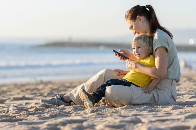 Mały blondyn i jego matka spędzają czas na plaży Kobieta patrzy na ekran telefonu