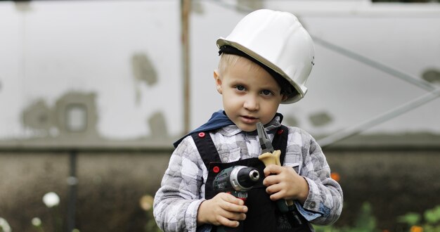 Mały budowniczy chłopiec w białym kasku siedzi na rusztowaniu ze szczypcami i wiertarką lub śrubokrętem