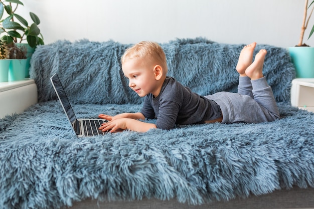 Mały Chłopiec Bawi Się Na Laptopie, Leżąc Na Kanapie W Domu. E-learning, Nauka Na Odległość, Koncepcja Komunikacji Na Odległość