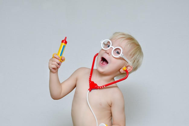 Mały chłopiec bawi się z lekarzem. Zabawkowa strzykawka, okulary i fonendoskop. Portret