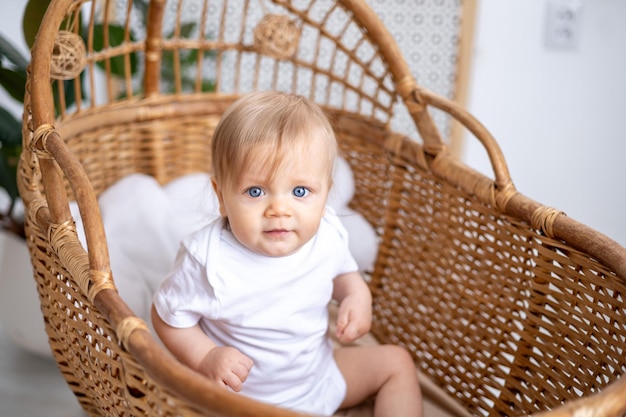 Zdjęcie mały chłopiec blondynka o niebieskich oczach w białym body siedzi w wiklinowej kołysce w domu w pokoju dziecinnym wnętrze pokoju dziecinnego