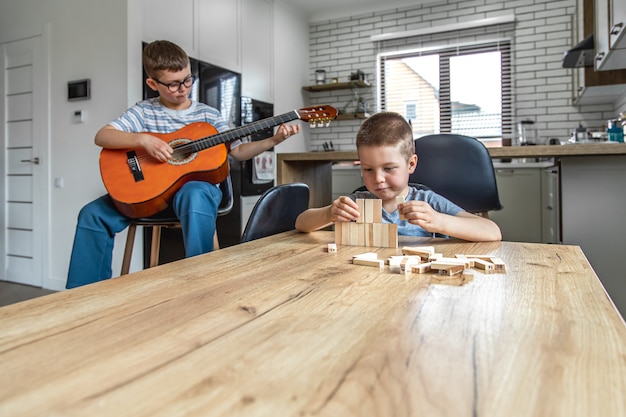 Mały Chłopiec Gra Na Gitarze, A Jego Brat Buduje W Domu Przy Stole Wieżyczkę Z Drewnianych Kostek.