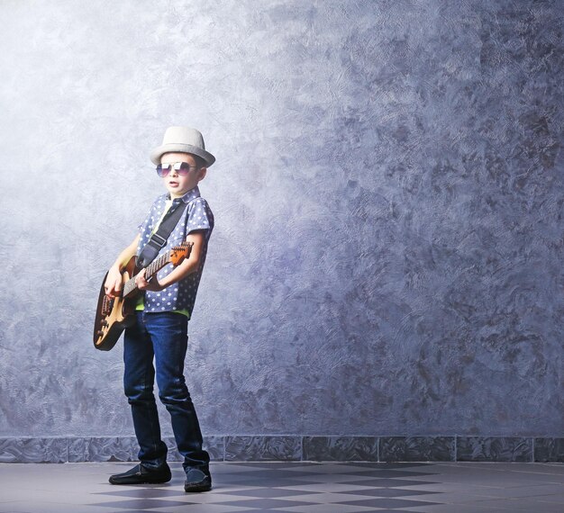 Mały chłopiec gra na gitarze na szarym tle ściany