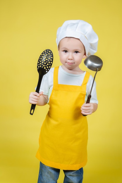 Mały chłopiec kaukaski grający w kucharza, chłopiec w fartuchu i czapce szefa kuchni pozuje na żółtym tle