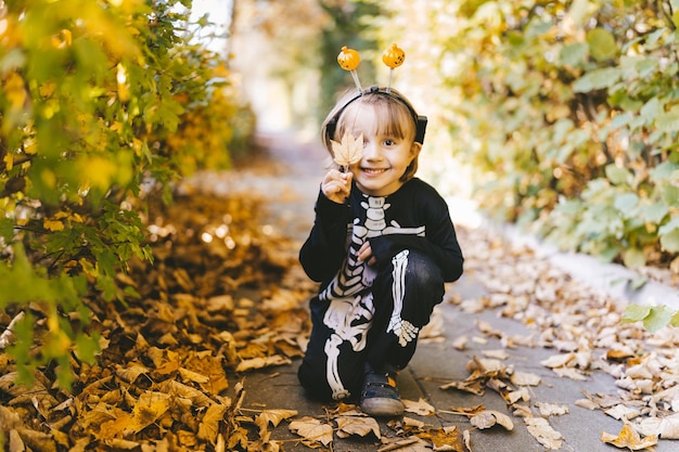 Mały chłopiec malucha w halloweenowym kostiumie szkieletowego karnawału siedzi na suchych liściach w parku jesienią Wszystkich Świętych Szkoła jesienne wakacje