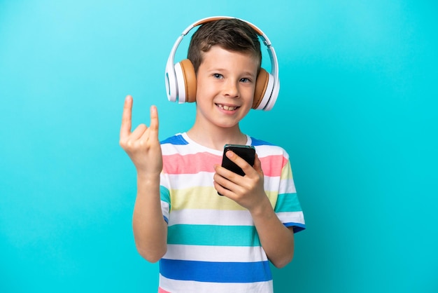 Mały chłopiec odizolowany na niebieskim tle słuchający muzyki za pomocą mobilnego gestu rockowego