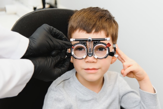 Mały Chłopiec Poważny Siedzi Na Krześle Gabinecie Badania Wzroku. Lekarz Podnosi Soczewki Do Specjalnych Okularów