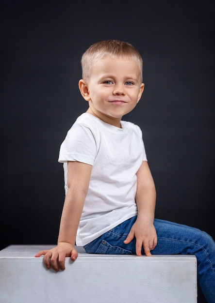 Mały chłopiec siedzi na białej kostce Uśmiechnięte dziecko w dżinsach i białej koszulce Czarne tło Pionowo