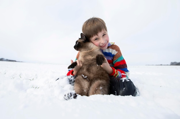 Mały chłopiec siedzi na śniegu i trzyma kota syjamskiego