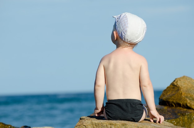 Mały chłopiec siedzi plecami do skały nad brzegiem morza w kąpielówki, błękitne niebo, miejsca na tekst
