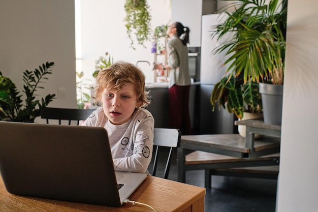 Mały chłopiec siedzi przy stole w kuchni i korzysta z laptopa, ucząc się online lub grając w grę