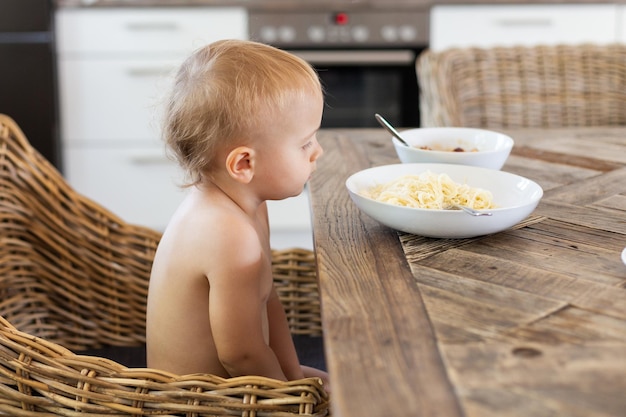 Mały chłopiec siedzi przy stole w kuchni i patrzy na talerz ze spaghetti z serem. Maluch jedzący obiad. Zdrowa żywność dla koncepcji dzieci.