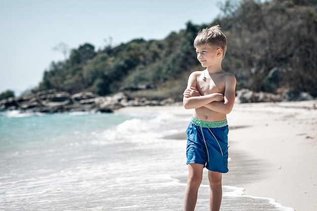 Mały chłopiec skrzyżował ramiona i stał w niebieskich spodenkach na piasku plaży. Mały chłopiec zabawy na tropikalnej plaży oceanu. Dziecko podczas rodzinnych wakacji na morzu. Letnia zabawa w wodzie. Phuket. Tajlandia