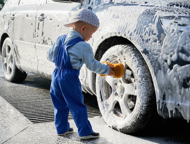 Mały chłopiec stojący w pobliżu samochodu pokryty pianką, trzymający gąbkę w dłoni i uczący się myć samochód Dziecko płci męskiej uczące się myć samochód gąbką pochyloną do przedniego koła