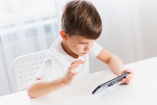 Mały chłopiec w białej koszulce gra w domu w gry przez telefon. Szczęśliwe dziecko patrzy na swój smartfon.