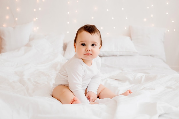 mały chłopiec w białych ubraniach uśmiecha się siedząc na białym łóżku