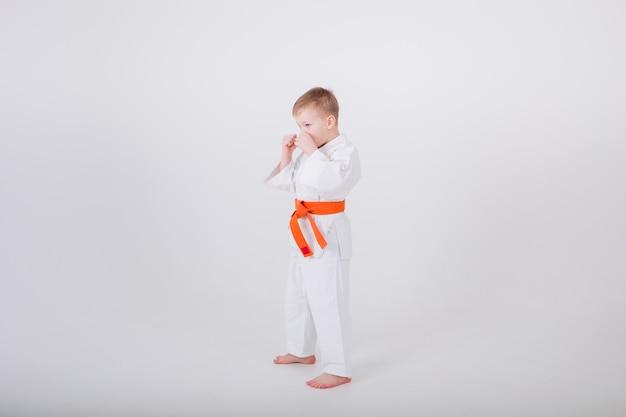 Mały chłopiec w białym kimonie z pomarańczowym paskiem stoi bokiem w ochronnej pozie na białej ścianie z kopią przestrzeni