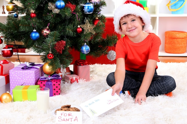 Mały chłopiec w czapce Świętego Mikołaja z mlekiem, ciastkami i listem do Świętego Mikołaja