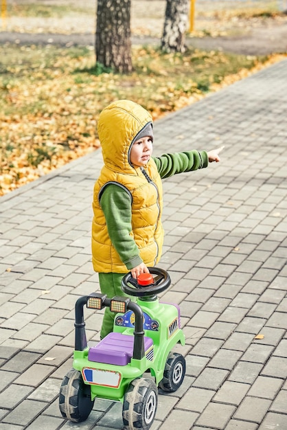 Mały chłopiec w jasnożółtej kamizelce z palcem stojącym w pobliżu zabawki Rideon idący po utwardzonej drodze