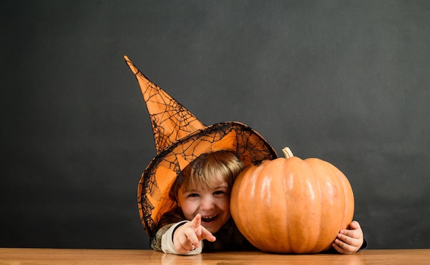 Mały chłopiec w kapeluszu wiedźmy z halloweenową dynią wskazujący na ciebie cukierek albo psikus przygotowujący na halloween