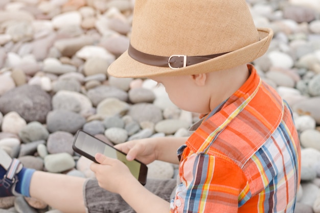 Mały Chłopiec W Kapeluszu Z Inteligentny Telefon Na Kamienistej Plaży