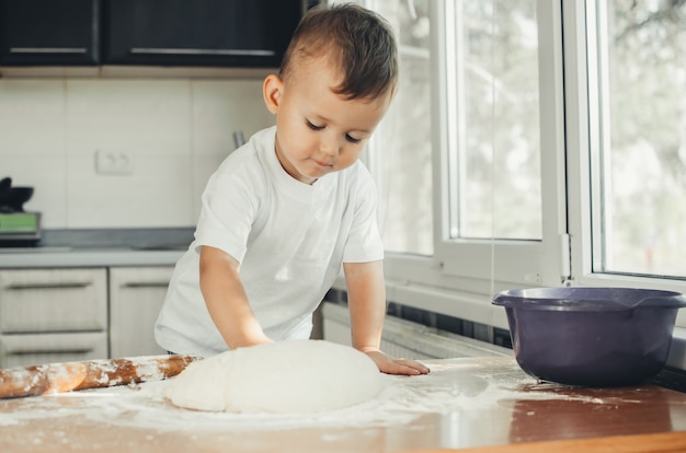 Mały Chłopiec w kuchni sam w białej koszulce mocno ugniata ciasto