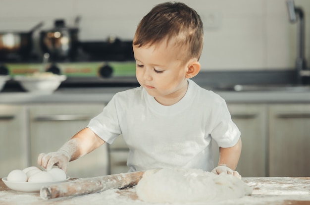 Mały Chłopiec w kuchni sam w białej koszulce mocno ugniata ciasto