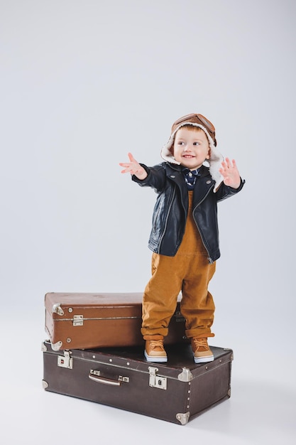 Mały chłopiec w skórzanej kurtce i brązowym kombinezonie stoi na dużej walizce Dziecko z walizką Mały podróżnik