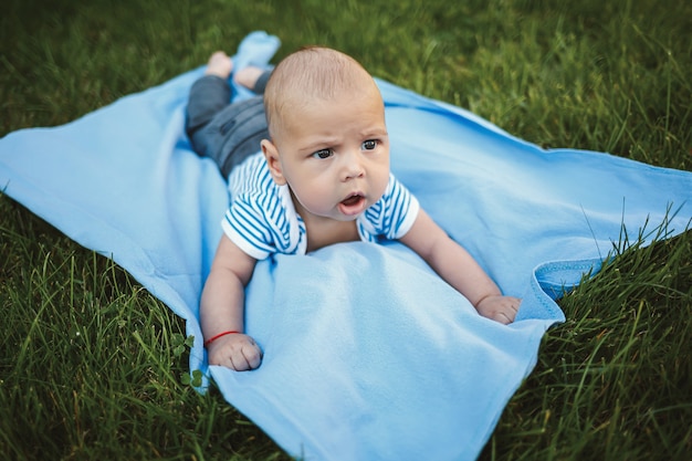 Mały chłopiec w wieku 3 miesięcy leży na brzuchu na niebieskiej narzucie w parku wokół zielonej trawy i drzew. Dziecięce emocje radości