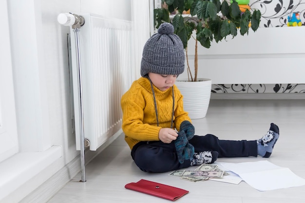 Mały chłopiec w żółtym swetrze i czapce liczy pieniądze i studiuje rachunki za ogrzewanie przy grzejniku z termostatem