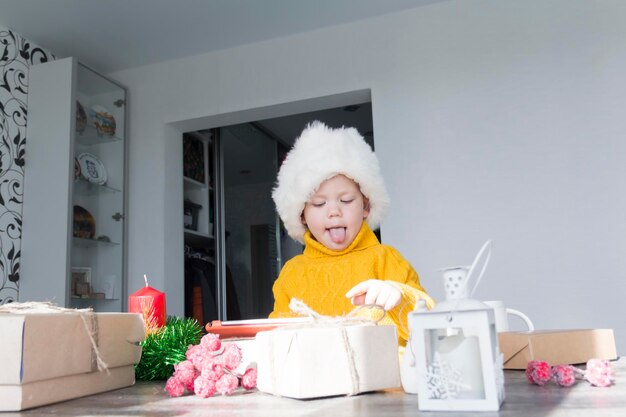 Mały chłopiec w żółtym swetrze i czerwonej czapce Świętego Mikołaja przy drewnianym stole pokazuje swój język