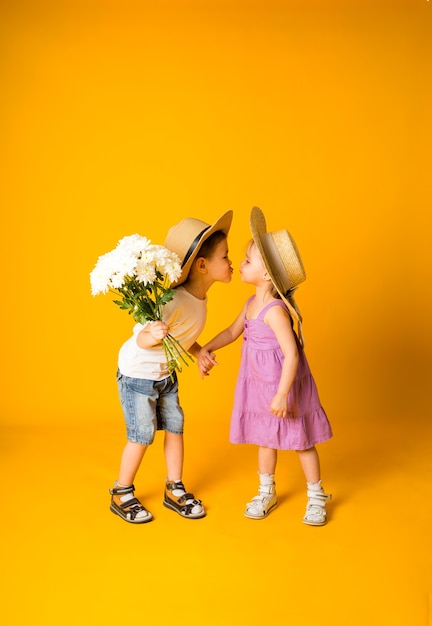 Mały chłopiec z bukietem kwiatów całuje małą dziewczynkę w słomkowym kapeluszu na żółtej powierzchni z miejscem na tekst