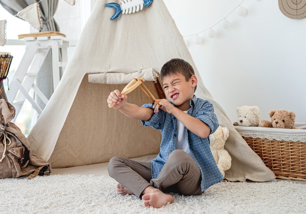 Mały Chłopiec Z Drewnianą Procą Siedzi Na Podłodze W Pokoju Przed Wigwamem