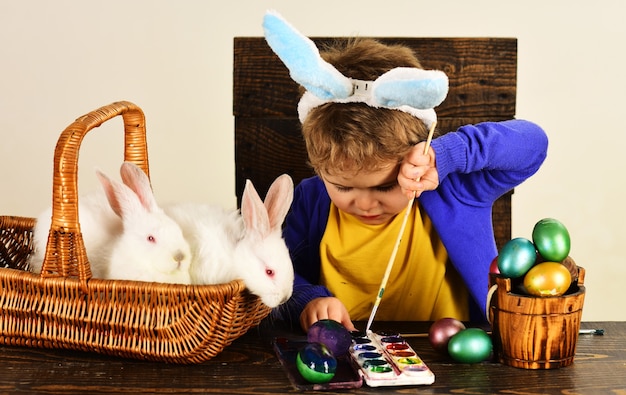 Mały chłopiec z Easter królikami w koszu. Dziecko w uszach królika maluje jajko wielkanocne.