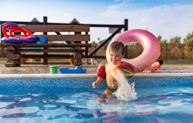 Mały chłopiec z jasnymi opaskami bawiącymi się kolorowymi zabawkami plażowymi w pobliżu ciemnoniebieskiego basenu z czystą przezroczystą wodą na tle ciepłego słonecznego letniego zachodu słońca Wideo 4K UHD slowmo