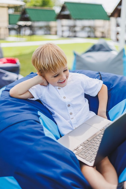 Mały chłopiec z laptopem na tle letniej trawy Chłopiec w białej koszulce siedzi na woreczku w parku