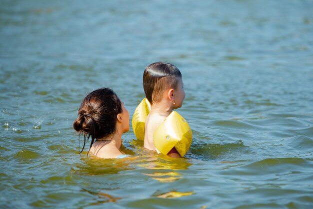 Mały chłopiec z nadmuchiwanymi opaskami pomaga bawić się w wodzie z matką w wodzie jeziora