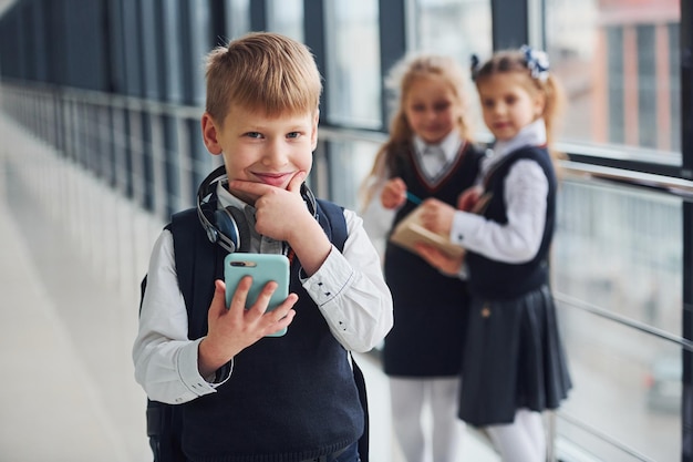 Mały chłopiec z telefonem i słuchawkami stojący przed dziećmi szkolnymi w mundurkach, które razem na korytarzu Koncepcja edukacji