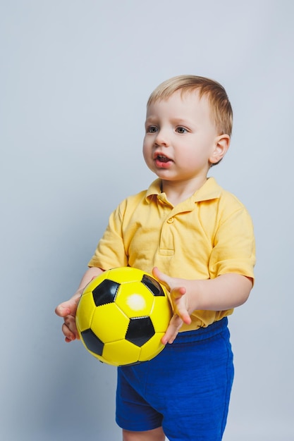 Mały europejski fan chłopiec lub gracz w żółtym i niebieskim mundurze z piłką nożną obsługuje drużynę piłkarską na białym tle Piłka nożna sport gra styl życia koncepcja izolowany na białym tle