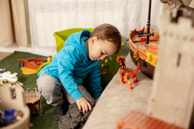 Mały kaukaski chłopczyk w niebieskiej bluzie bawi się kolorowymi zabawkami w domu Zabawa dziecka Wesoły i wesoły dzieciak bawi się statkiem Dinozaurowy zamek Aktywność w czasie wolnym życie domowe Przytulny pokój