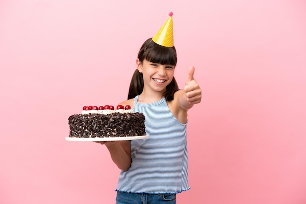 Mały kaukaski dzieciak trzymający tort urodzinowy na białym tle na różowym tle z kciukami do góry, ponieważ stało się coś dobrego