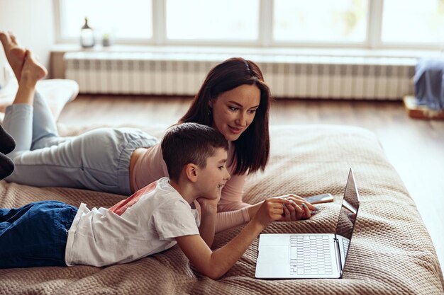 Mały kaukaski ładny chłopiec z piękną matką leżącą razem na łóżku z laptopem