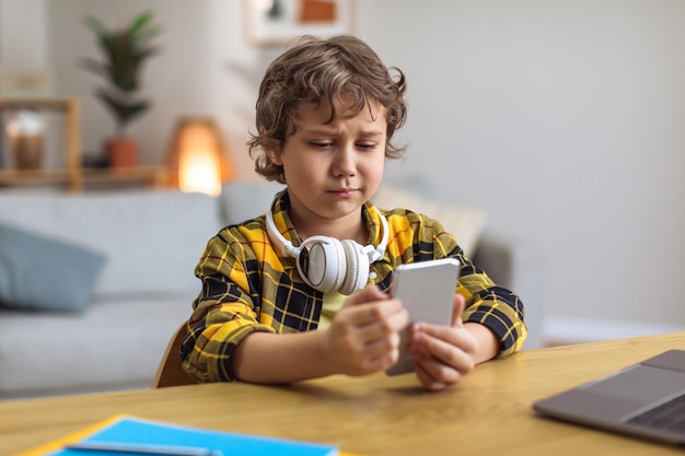 Mały niezadowolony chłopiec patrzący ze smutkiem na smartfona, przegrywający grę wideo lub czytający nieprzyjemną wiadomość, denerwujący