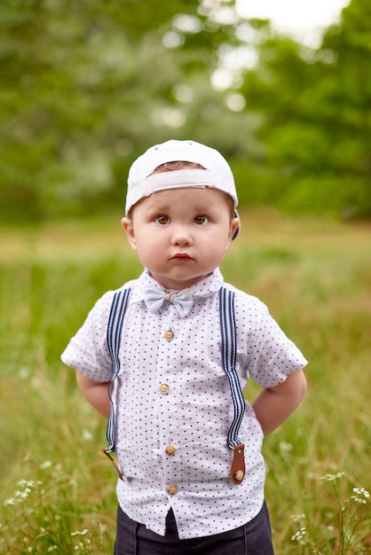 Zdjęcie mały poważny chłopiec w szelkach i czapce chowa ręce za plecami w parku
